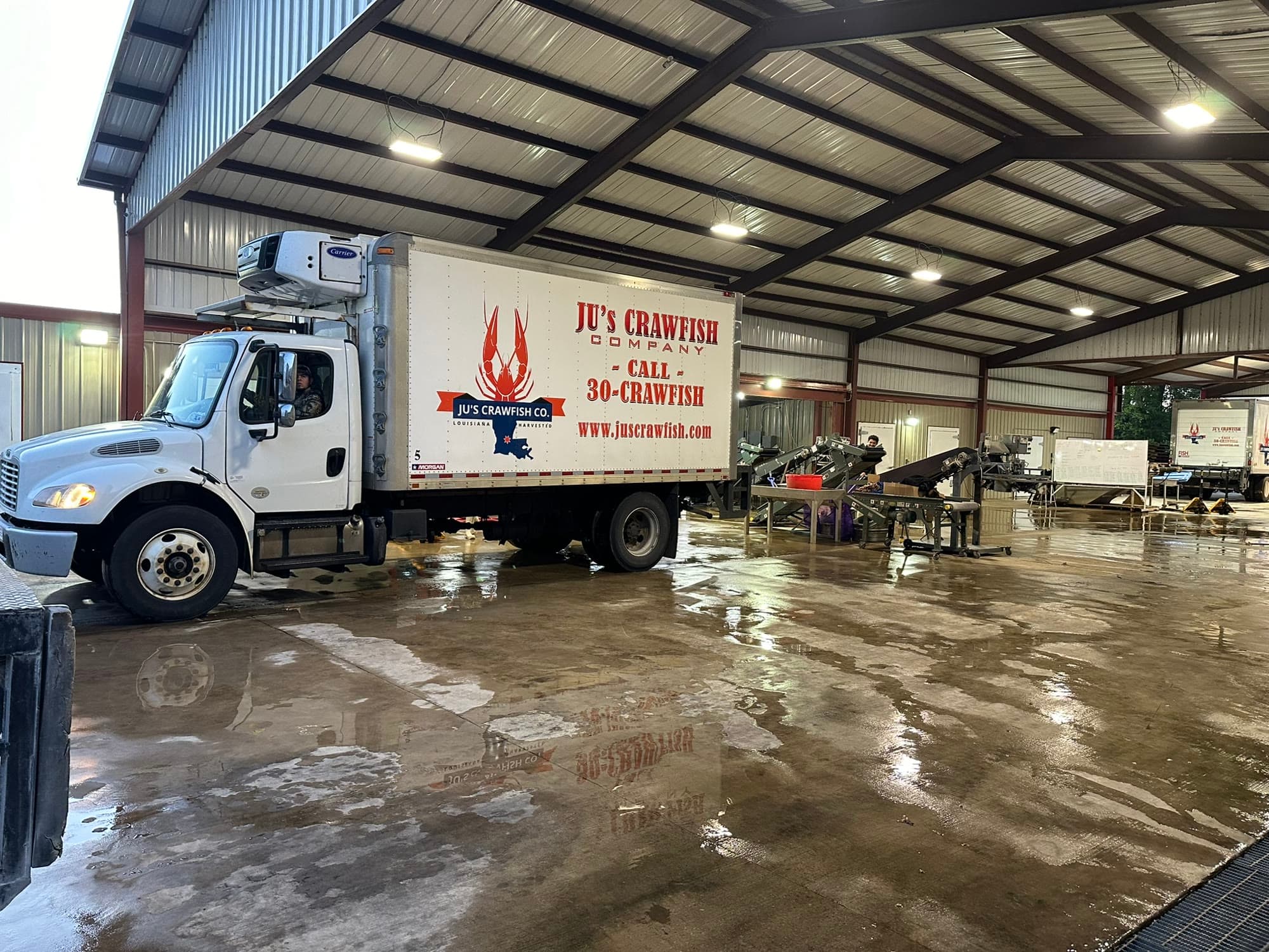 A branded Ju's Crawfish Company refrigerated truck parked inside the Plaucheville plant with the wash-and-grade line running along the back wall